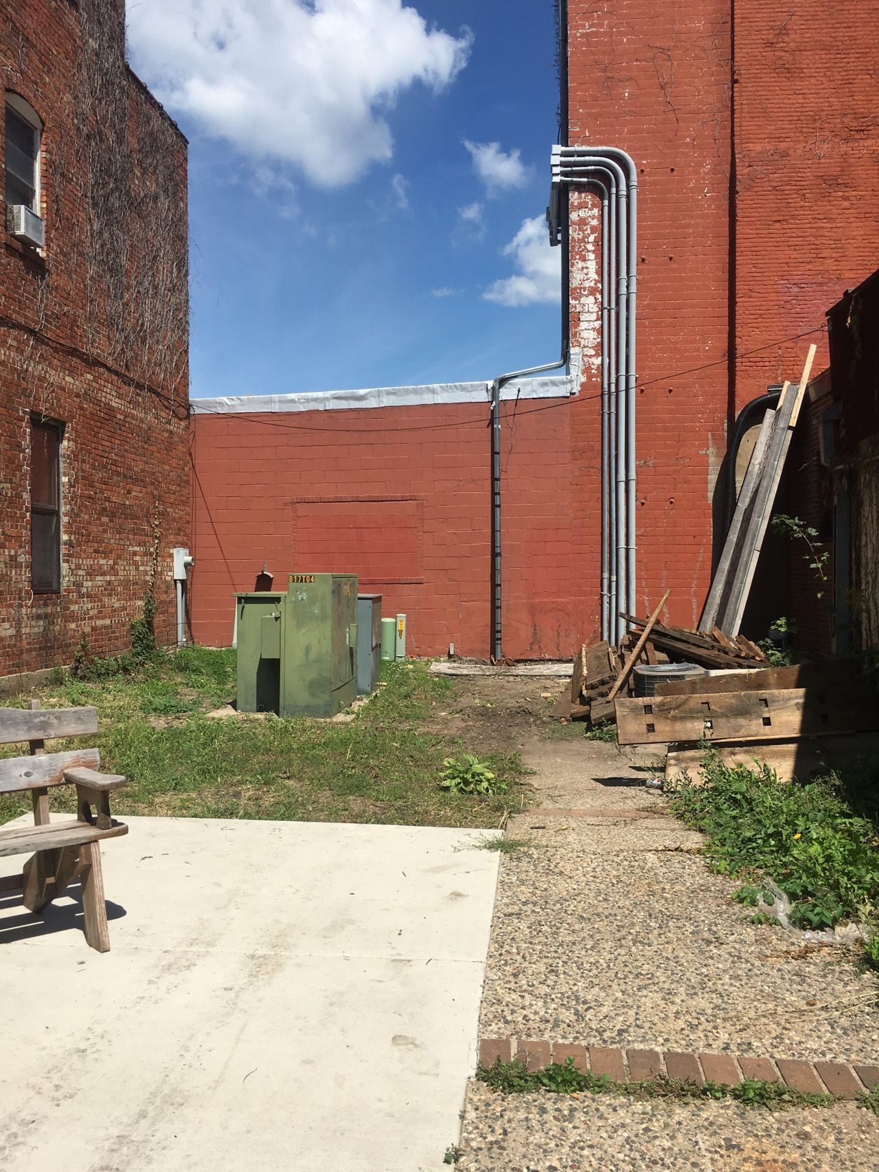 The empty back lot today, with utility boxes, scrap lumber, and patchy grass between brick walls